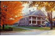 A large, multi-story stone building with a covered porch and white columns, surrounded by trees with vibrant orange autumn leaves, situated next to a paved road.