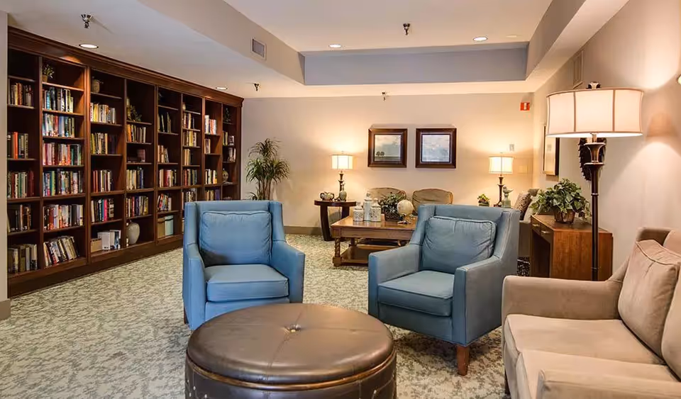 A cozy senior living common area with two blue armchairs, a beige sofa, a round brown ottoman, and a large wooden bookshelf filled with books. The room is softly lit with table lamps and a floor lamp, and there are framed pictures on the wall and a small plant on a side table.