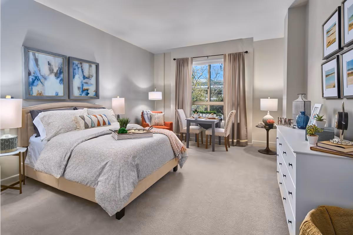 A bright and cozy bedroom with a large bed dressed in light-colored bedding, two abstract paintings above the headboard, bedside tables with lamps, an orange armchair with a throw blanket, a small dining table with two chairs near a window with beige curtains, and a white dresser decorated with plants, books, and framed pictures.