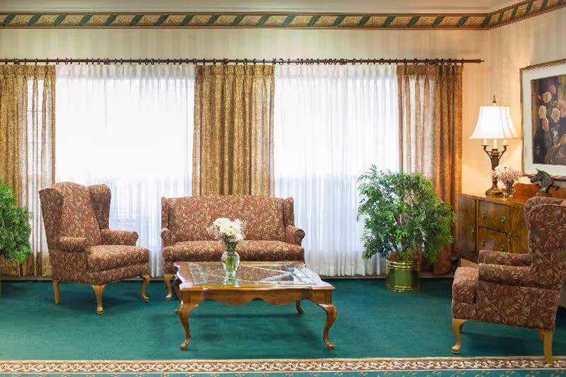 Carpeted seating area with patterned upholstered sofa and chairs around a wooden coffee table in front of large curtained windows.