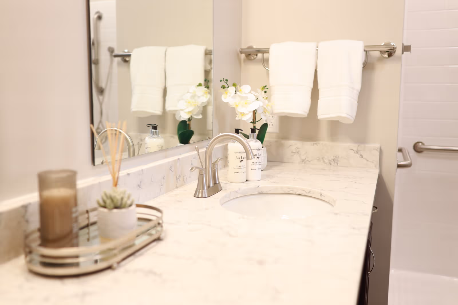 A clean bathroom countertop with a built-in sink and silver faucet. On the countertop, there is a small tray holding a candle, a small potted plant, and reed diffuser sticks. A bottle of hand lotion and a white orchid plant are placed near the sink. Two white towels hang on a silver towel rack above the counter. The background shows a tiled shower area with a grab bar.
