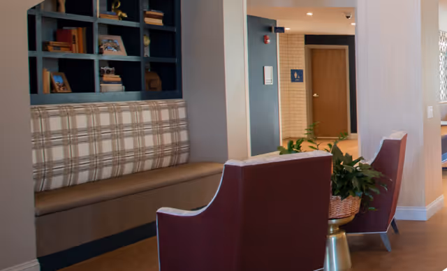 Interior view of a senior living facility lounge area with a built-in plaid cushioned bench, two maroon armchairs with white trim, a small round table with a potted plant, and a bookshelf with decorative items. In the background, there is a hallway with a restroom door and signage.