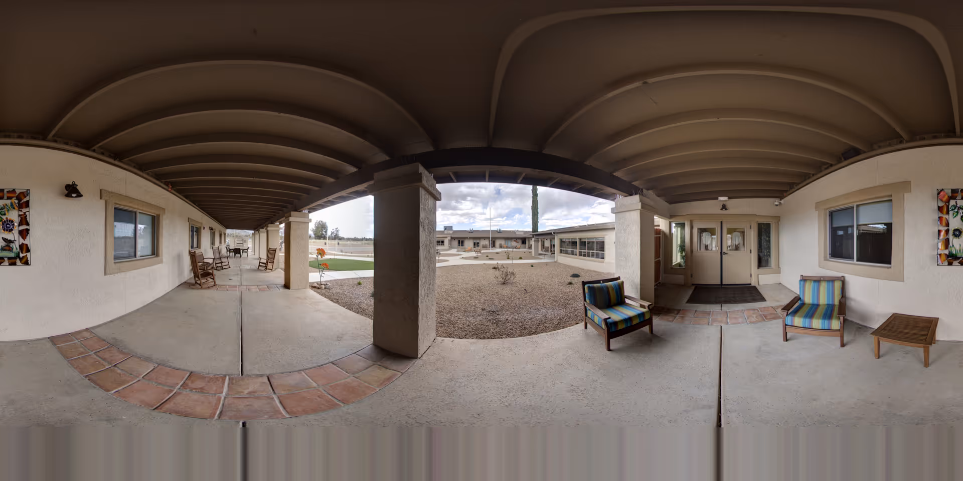 Covered outdoor walkway with tiled floor sections and seating including rocking chairs and cushioned chairs with a small table. The walkway surrounds a central courtyard area with gravel and sparse desert landscaping. The building exterior is light-colored with windows and decorative wall art. The sky is partly cloudy.