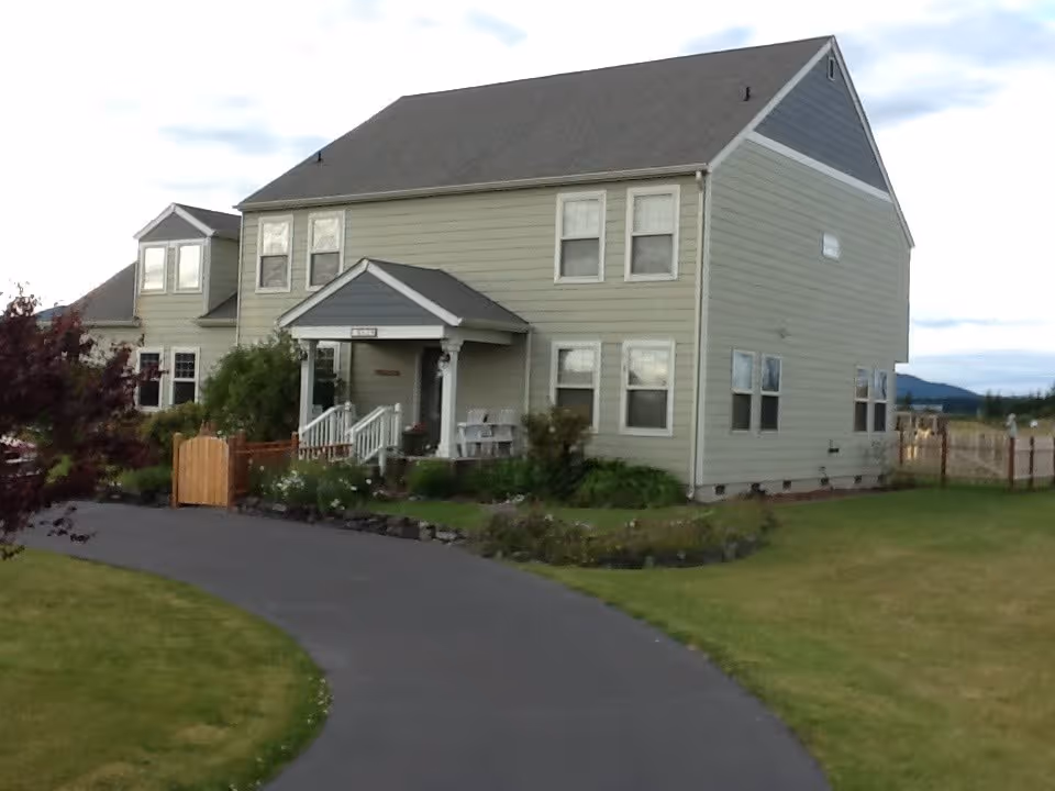 Two-story pale green house with a front porch, curved driveway, and surrounding lawn and landscaping.