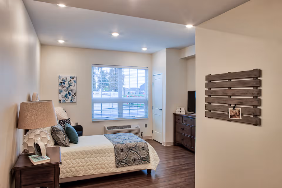 A cozy bedroom in Demaree Crossing featuring a neatly made bed with white quilted bedding and decorative pillows. A wooden nightstand with a lamp and books is beside the bed. The room has a large window with blinds, a wooden dresser with a TV on top, and a decorative wooden wall piece with a photo clipped to it. The floor is wooden, and the walls are painted light beige.