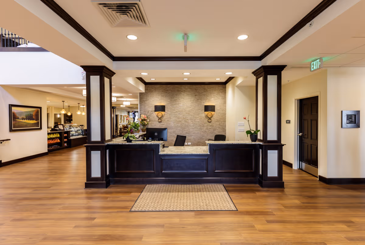 Reception desk area in a senior living facility with dark wood paneling and a granite countertop. The background wall has a textured stone finish with two wall sconces. The floor is wood with a small rug in front of the desk. To the left, there is a hallway leading to a dining area with hanging lights and a display case. An exit sign is visible on the ceiling to the right.