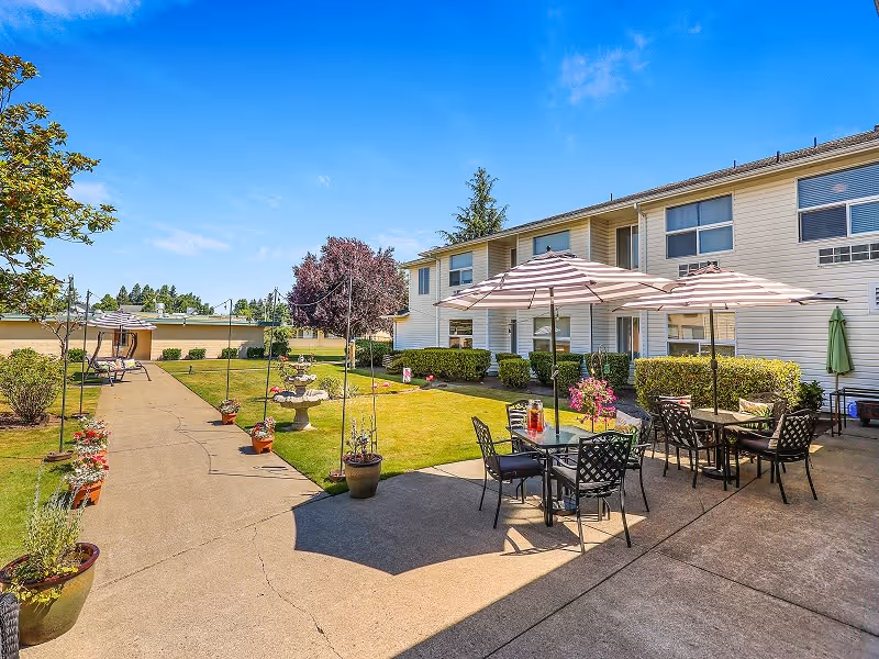 Outdoor patio area at White River Assisted Living with tables and chairs under striped umbrellas, surrounded by potted plants and greenery, adjacent to a two-story building under a clear blue sky.