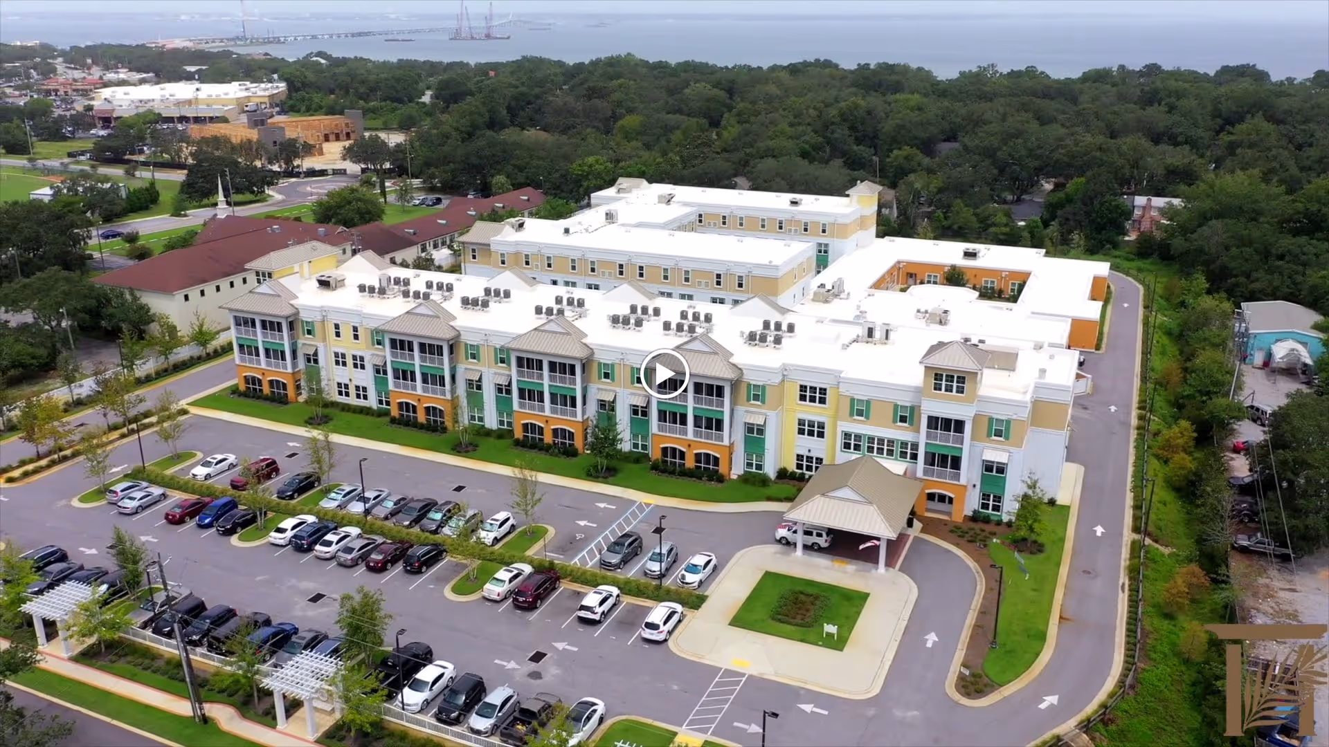 Aerial view of The Arbors of Gulf Breeze senior living facility showing a large multi-story building with white roofs and pastel-colored walls, surrounded by parking lots with numerous cars, landscaped greenery, and trees in the background.