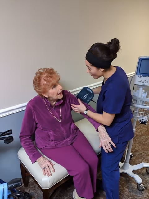 A healthcare worker in navy scrubs is taking the blood pressure of an elderly woman wearing a purple outfit, who is seated on a cushioned bench in a medical or supportive living facility room.