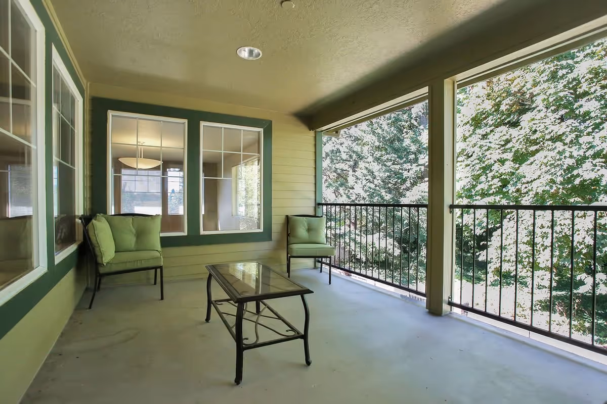 A covered outdoor balcony area with two green cushioned chairs and a glass-top coffee table. The balcony has a railing and overlooks trees and greenery outside. The walls are painted green with white trim around the windows.