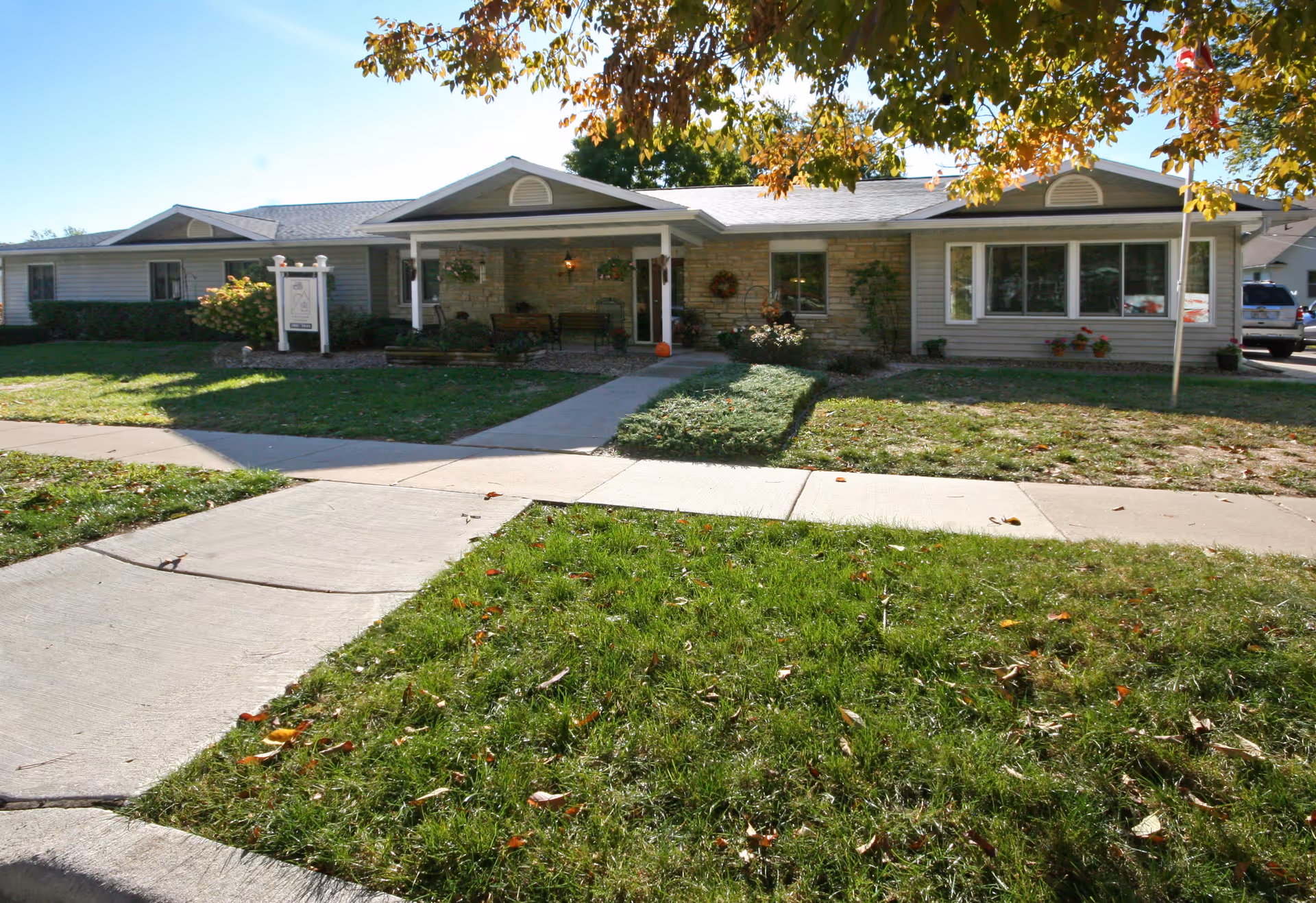 Exterior front view of a single-story senior living facility building with a covered porch, stone and siding facade, surrounded by grass and trees with autumn leaves.