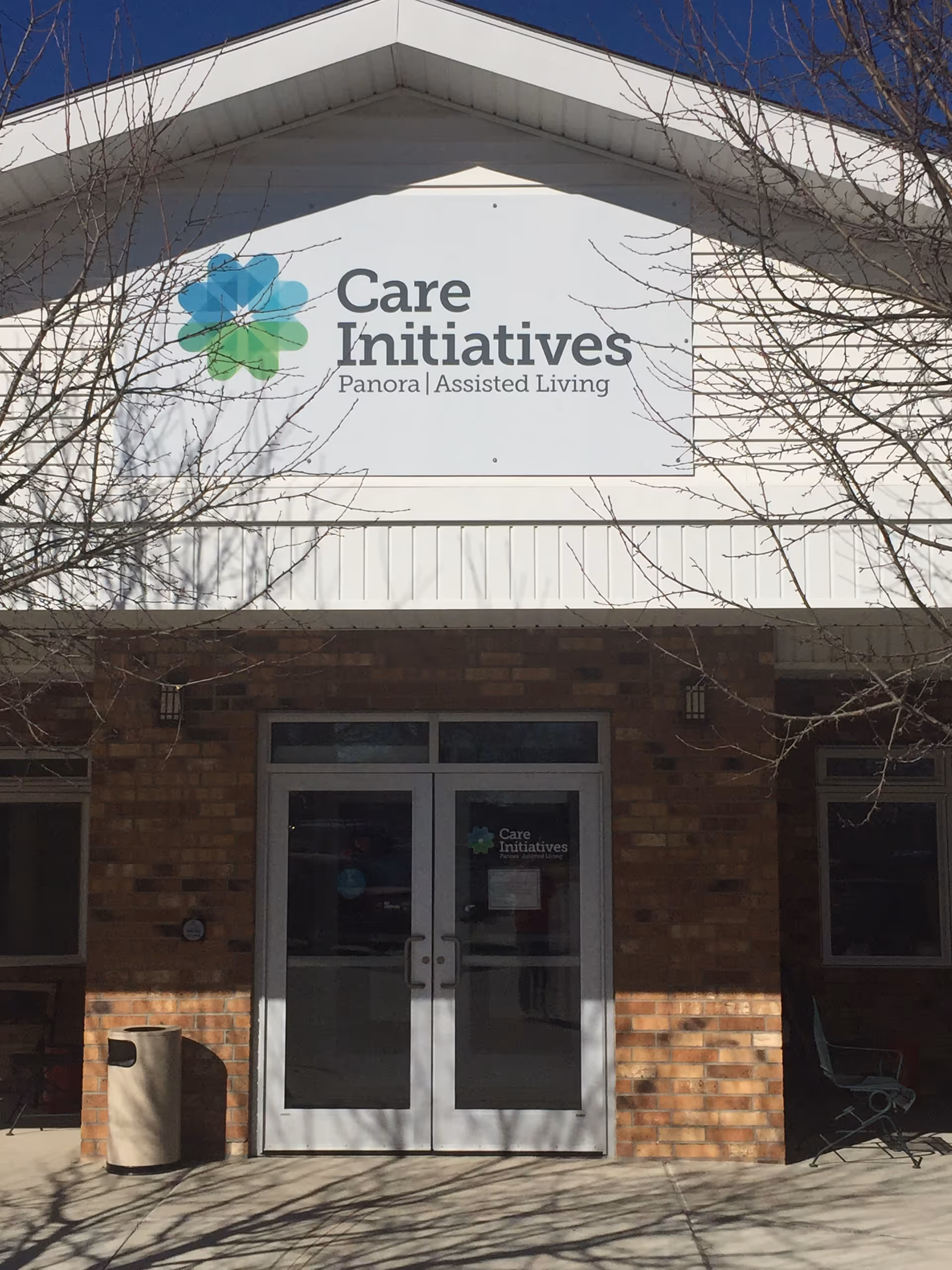 Front entrance of Care Initiatives Panora Assisted Living facility with double glass doors, a brick exterior, and a large sign above the entrance displaying the facility name and logo. Leafless tree branches are visible in the foreground.