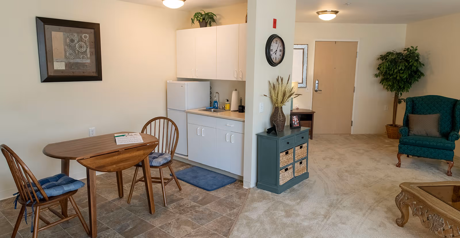 Interior view of a senior living facility unit at Creekside Terrace showing a small kitchenette with white cabinets, a refrigerator, and a sink. Adjacent to the kitchenette is a wooden dining table with two chairs, each with blue cushions. The living area has a green upholstered armchair, a wooden coffee table, a small blue cabinet with wicker baskets, a wall clock, and a potted plant near the entrance door.