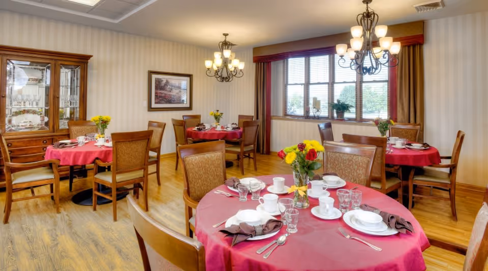 A dining room with round tables covered in red tablecloths, each set with white plates, cups, glasses, and silverware. Each table has a vase with yellow and red flowers. Wooden chairs surround the tables. The room has wooden flooring, a large window with curtains, two chandeliers, a wooden cabinet with glass doors displaying dishes, and a framed picture on the wall.