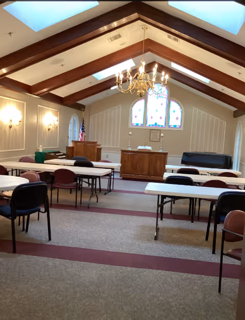 Interior view of a chapel or meeting room with vaulted ceiling and wooden beams, featuring a chandelier, stained glass windows, an American flag, a wooden podium, and several tables and chairs arranged in rows.