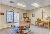 A small room with a round wooden table surrounded by four folding chairs. The room has beige walls and carpeted floor. On one wall, there is a large cross, a table with a lamp, flowers, and framed pictures. Another wall features a stained glass window with colorful designs and a clock above it. The ceiling has fluorescent lights.