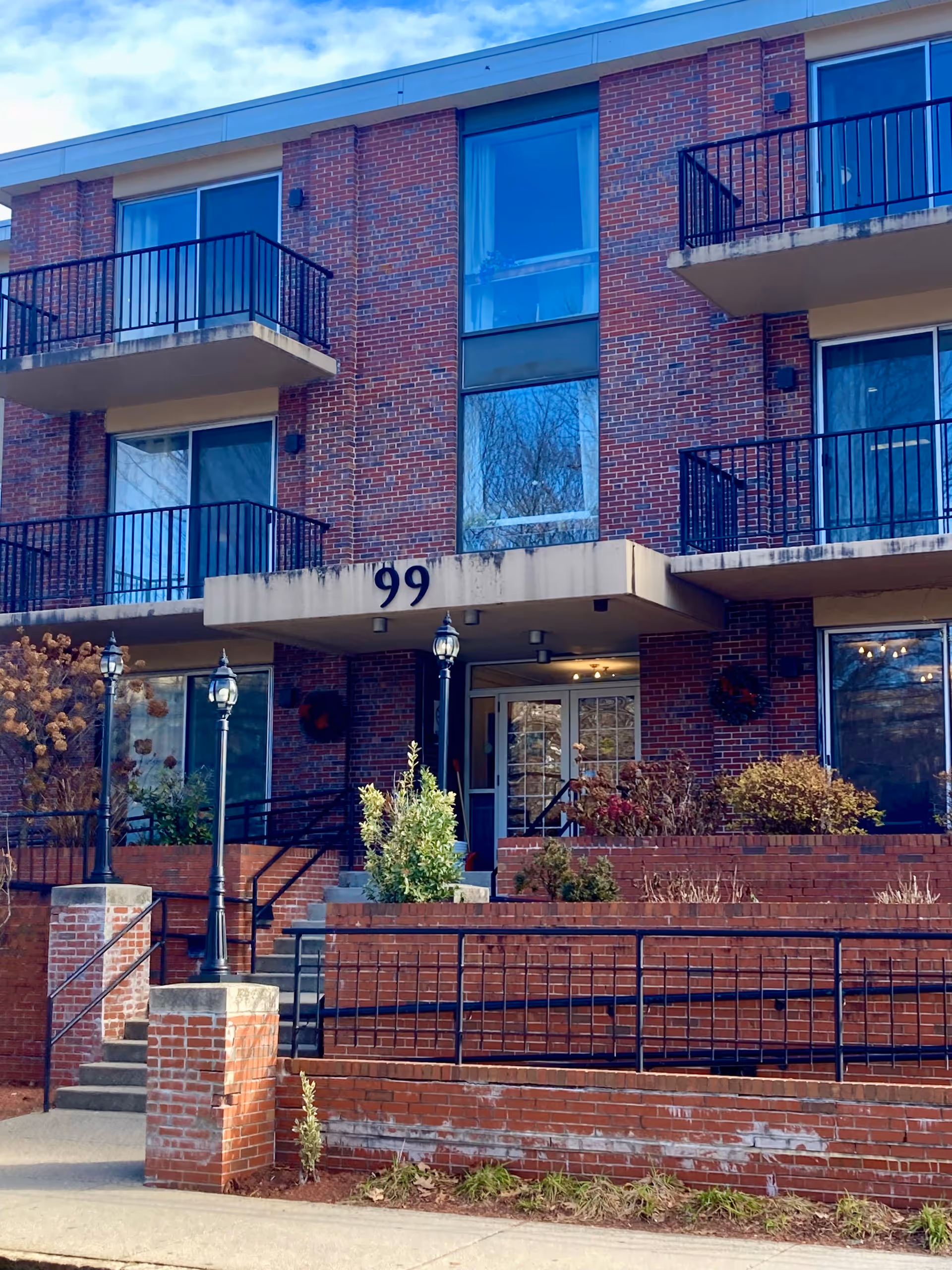Exterior view of a brick building with balconies and large windows. The entrance is marked with the number 99 above a covered porch. There are stairs and black metal railings leading up to the entrance, with some landscaping including bushes and lamp posts.