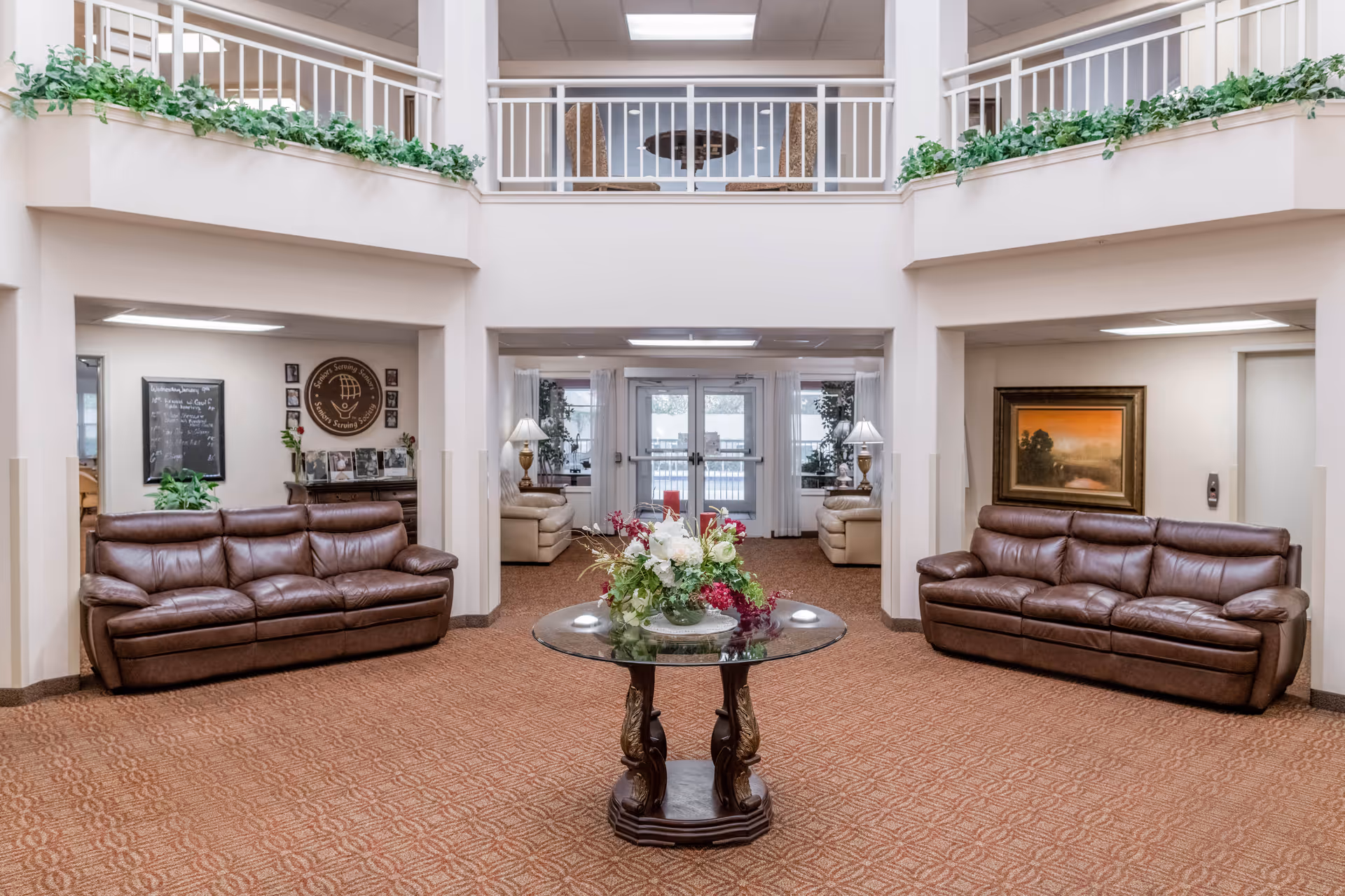 A spacious and well-lit common area with two brown leather sofas facing each other, a round glass table with a floral arrangement in the center, and a second-floor balcony with white railings and green plants. The room has beige walls, carpeted floor, and large windows with white curtains letting in natural light.