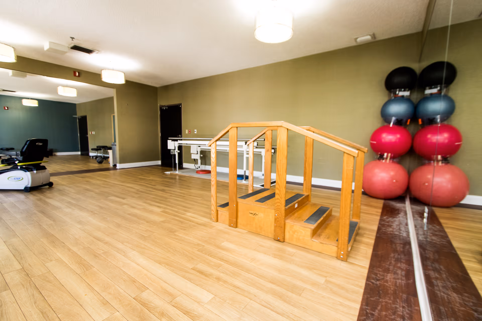 A spacious rehabilitation or exercise room with wooden flooring, a small wooden staircase with handrails, exercise balls stacked in the corner, a recumbent exercise bike, and parallel bars for physical therapy. The room has green walls and ceiling lights.