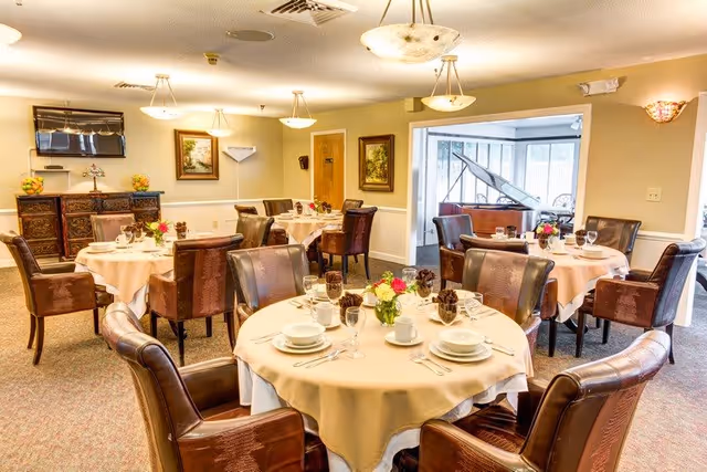 Well-lit dining room with round tables set for service, leather chairs, floral centerpieces and a grand piano in the background.