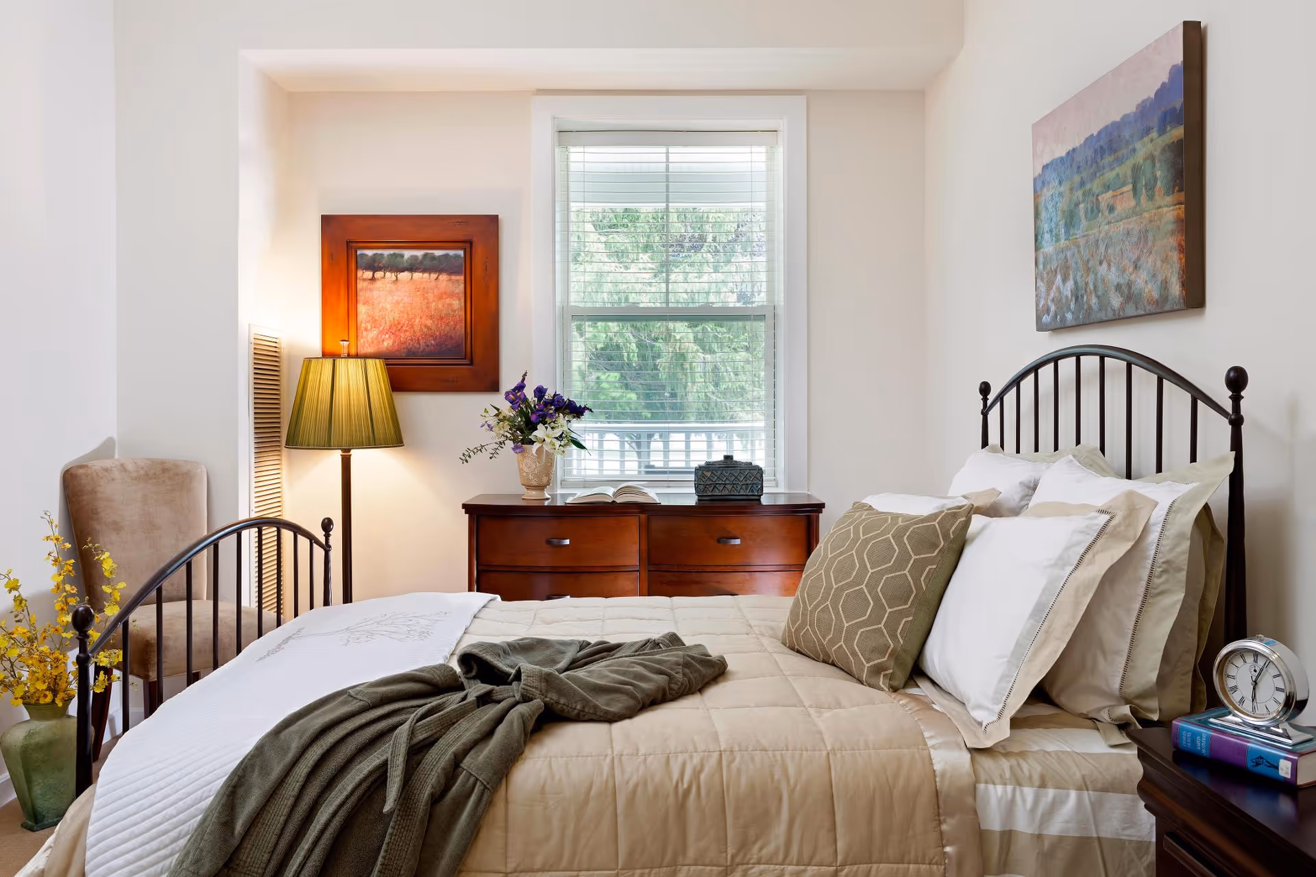 Sunlit bedroom with a made bed featuring decorative pillows and a metal headboard, a dresser under a window, a lamp, and wall artwork.