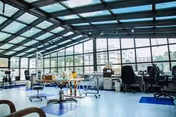 A spacious rehabilitation therapy room with large floor-to-ceiling windows and a glass ceiling allowing natural light to fill the space. The room contains various physical therapy equipment including parallel bars, exercise machines, chairs, and tables. The floor is covered with blue linoleum, and the overall atmosphere is bright and airy.