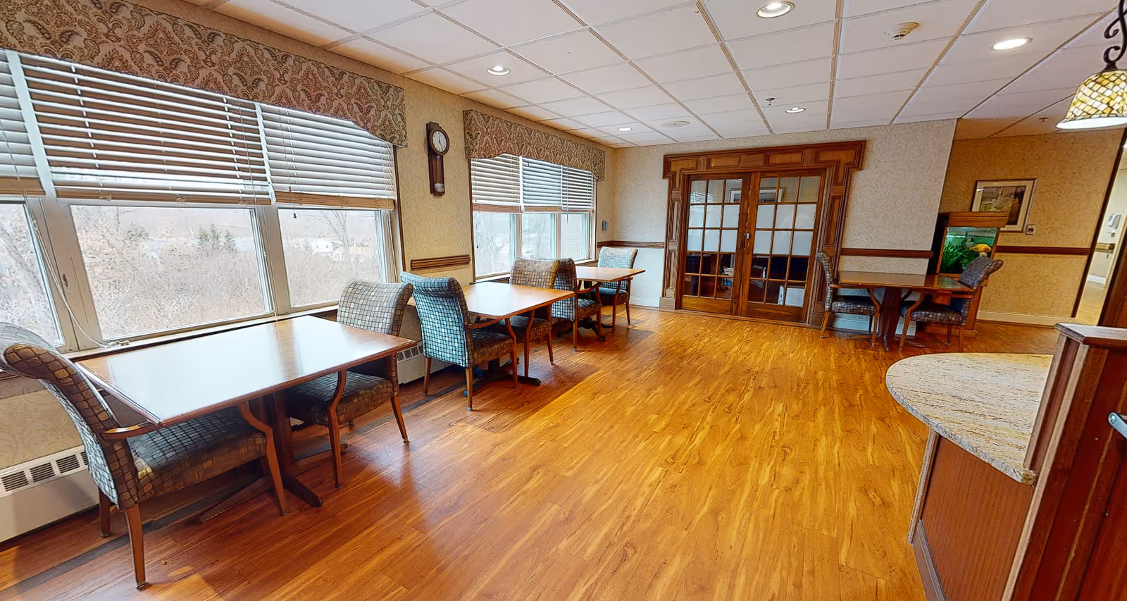 A bright dining area with wooden floors and several tables, each accompanied by two upholstered chairs. Large windows with blinds let in natural light, and a wall clock is mounted between the windows. The room features a set of wooden double doors with glass panels and a small fish tank on a side table near the entrance.