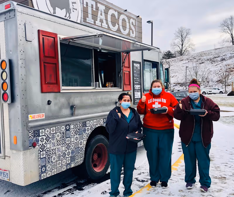 Three people wearing face masks and scrubs stand outside in the snow next to a taco food truck. They are holding food trays and giving thumbs up. The food truck has a sign that says TACOS and decorative tiles on the lower half.