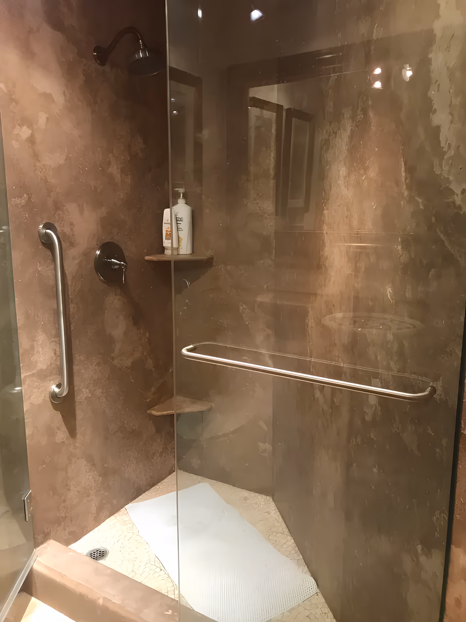 A modern shower area with brown textured walls, a glass door, a metal grab bar on the wall, a showerhead, and two corner shelves holding bottles of soap or shampoo. There is a white mat on the floor inside the shower.