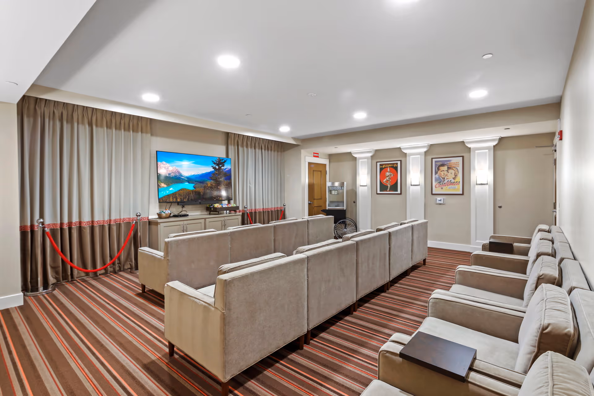 A cozy media room with rows of beige seats facing a large wall-mounted TV, striped carpet, and framed posters on the far wall.