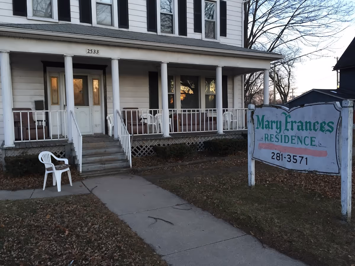 Exterior view of a two-story white building with a porch featuring several chairs. A sign in front reads 'Mary Frances Residence' with a phone number below. The building has multiple windows and a central door with steps leading up to the porch.