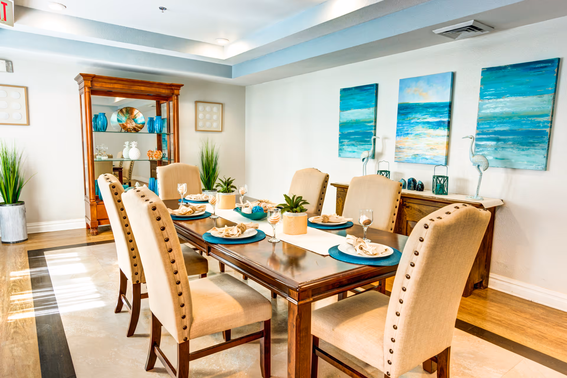 A bright dining room with a wooden table set for six people. The table has beige upholstered chairs with nailhead trim, blue placemats, beige napkins, and wine glasses. The room features wooden flooring with a light-colored rug under the table, a wooden cabinet with decorative items, and three blue and green ocean-themed paintings on the wall. There are also two tall plants in metallic pots and a wooden display cabinet with glass shelves holding decorative items.