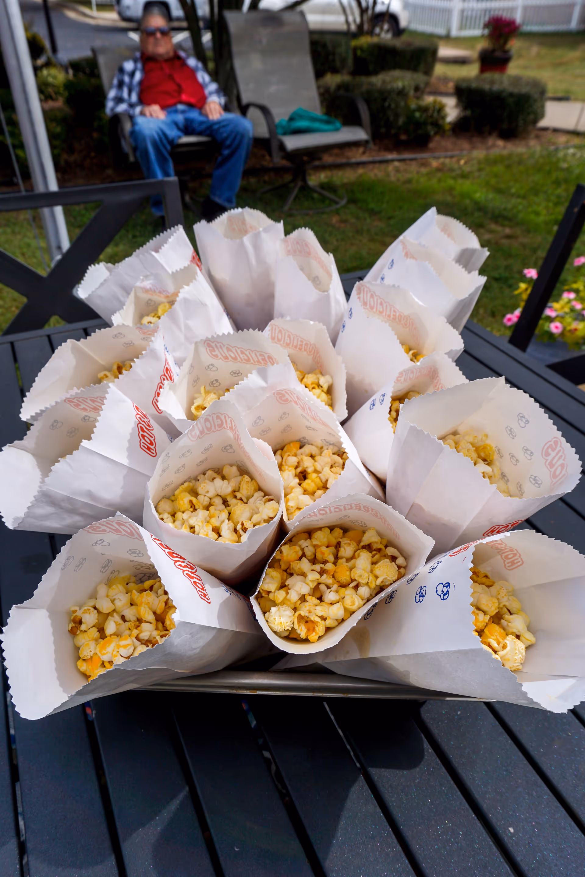 Multiple paper bags filled with popcorn arranged on a tray on an outdoor table. In the background, an elderly person is sitting on a chair in a garden area with greenery and flowers.