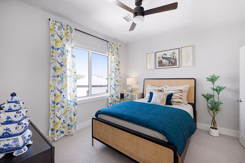 A bright and cozy bedroom with a bed featuring a woven headboard and footboard, dressed in white and teal bedding with decorative pillows. The room has a window with floral yellow and blue curtains, a ceiling fan, a nightstand with a lamp and picture frame, a potted plant in the corner, and blue and white decorative ceramic jars on a dark dresser.