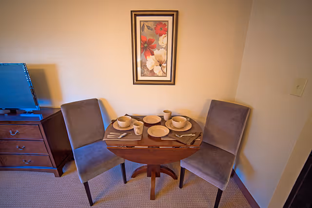 A small dining area with a round wooden table set for two, including plates, bowls, cups, and utensils. Two upholstered chairs are positioned on either side of the table. A framed floral painting hangs on the wall above the table. To the left, there is a wooden dresser with a flat-screen TV on top.