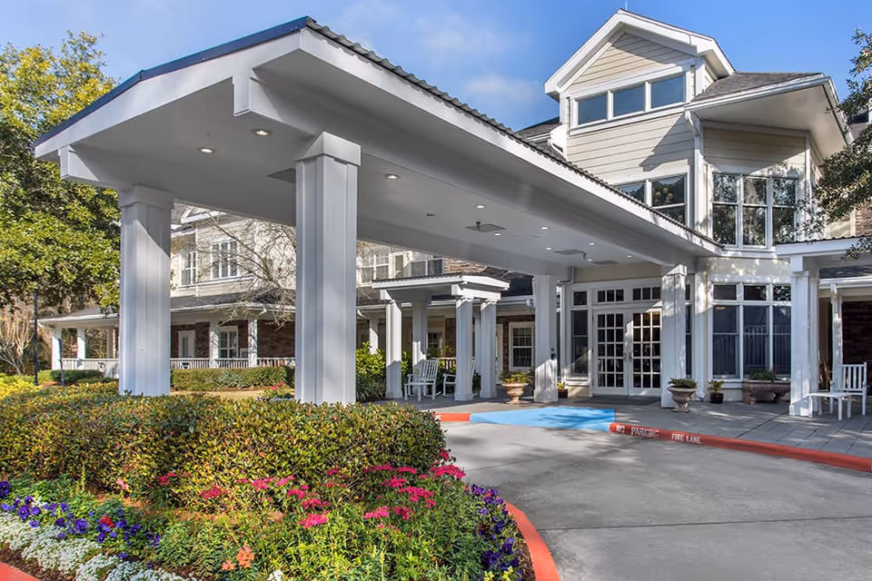 Entrance of a senior living facility with a covered drop-off area supported by white pillars, surrounded by well-maintained landscaping with colorful flowers and greenery. The building has large windows and a welcoming appearance under a clear blue sky.