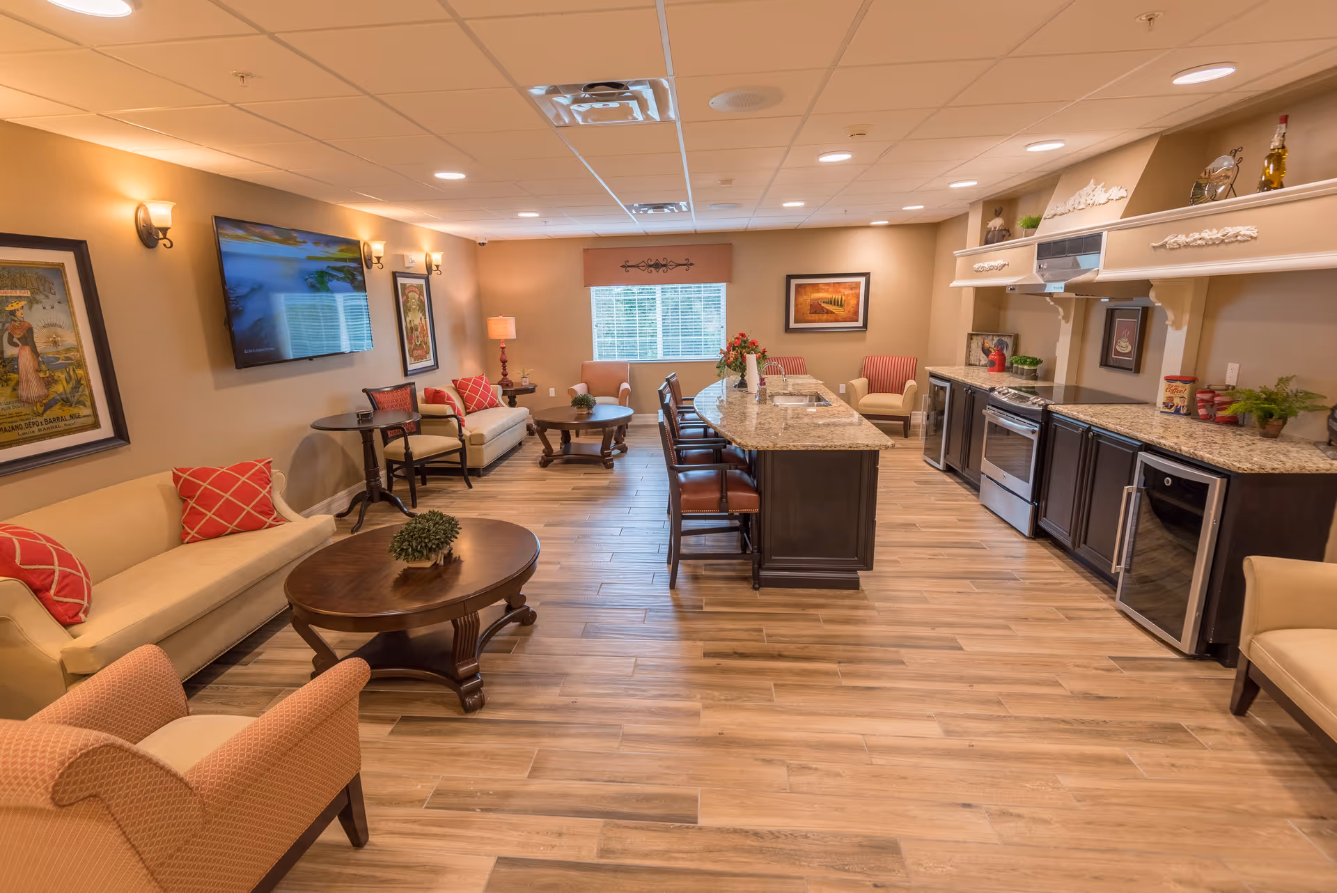 A spacious and well-lit common area featuring a kitchen island with bar stools, granite countertops, and modern appliances on the right. The left side has comfortable seating including sofas and armchairs with red and beige cushions. The walls are decorated with framed artwork and a mounted flat-screen TV. The room has wooden flooring and a large window at the back letting in natural light.