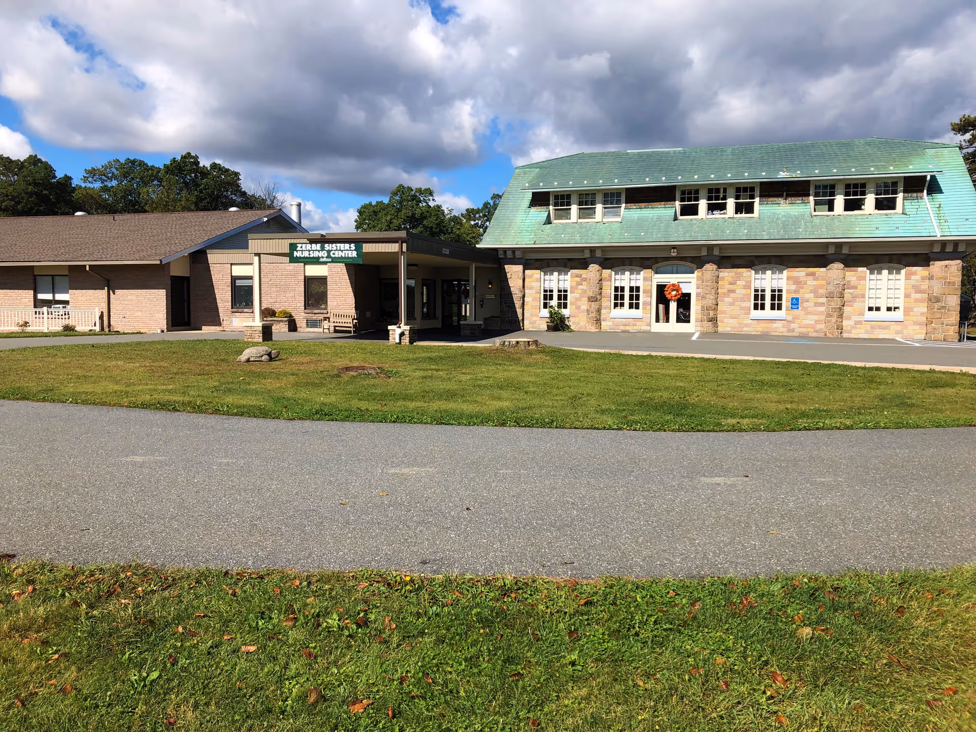 Exterior view of Zerbe Sisters Nursing Center, part of Zerbe Retirement Community, showing a building with a green roof and stone columns, a covered entrance, and a grassy area in front under a partly cloudy sky.