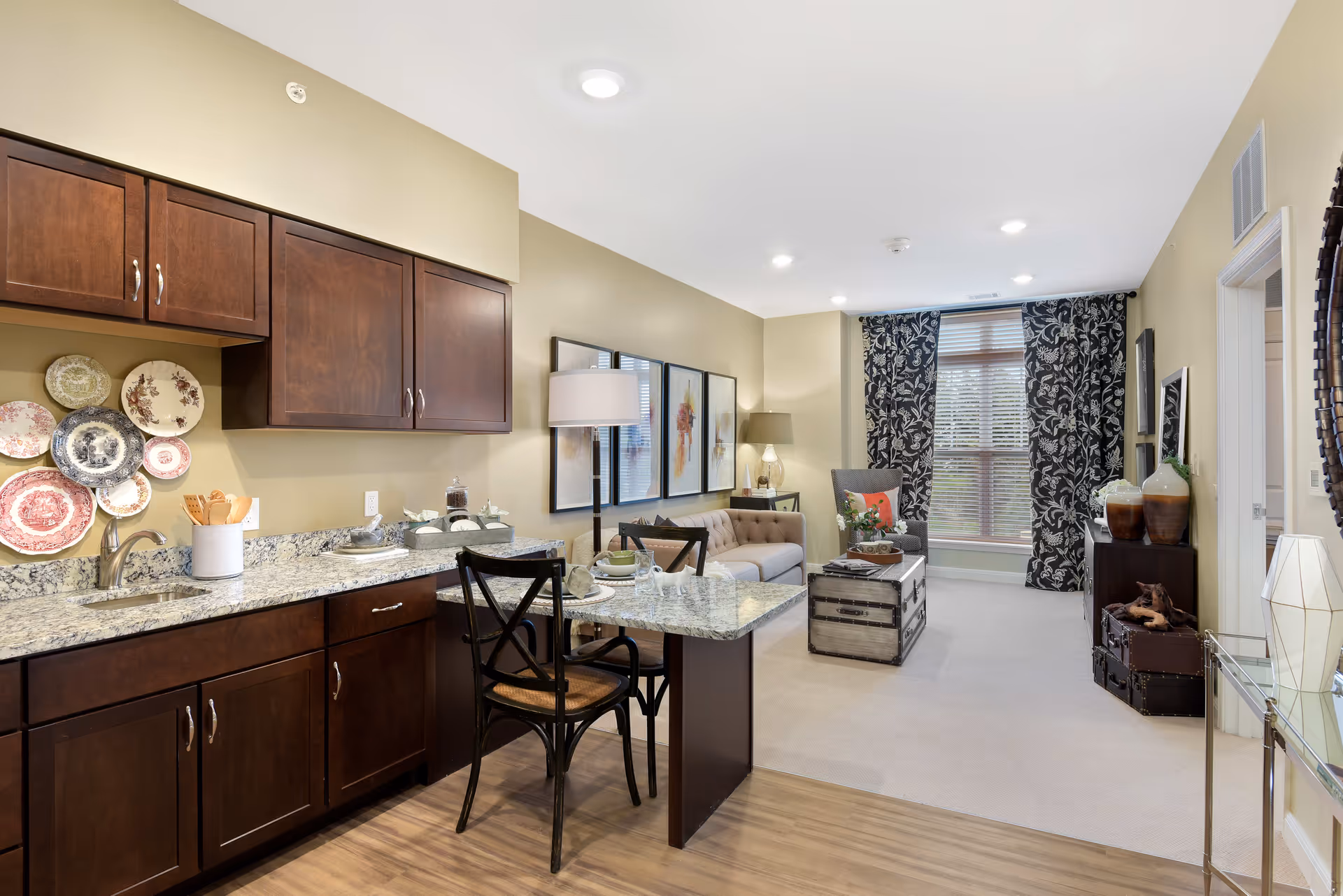 A cozy living space featuring a kitchen area with dark wooden cabinets and granite countertops on the left. The kitchen counter extends into a small dining area with two black chairs. The living room area has a beige sofa, a gray armchair, a wooden chest used as a coffee table, decorative plates on the kitchen wall, and large windows with patterned black and white curtains letting in natural light.