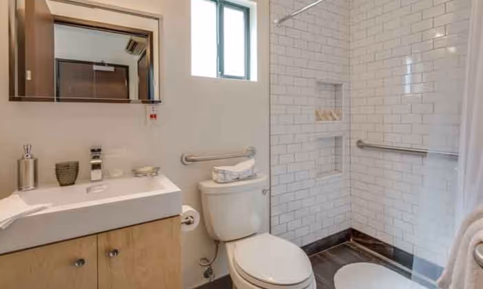 A clean and modern bathroom featuring a white toilet with a towel on top, a white sink with a wooden cabinet below, a mirror above the sink, and a shower area with white subway tiles and built-in shelves. There are grab bars installed near the toilet and inside the shower for accessibility.