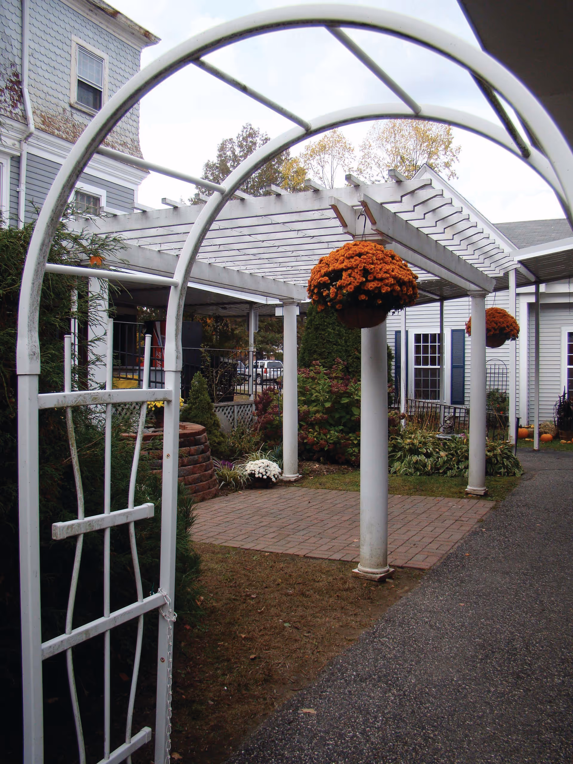 White pergola-covered courtyard with hanging orange flower baskets, columns, and a brick patio beside a building.