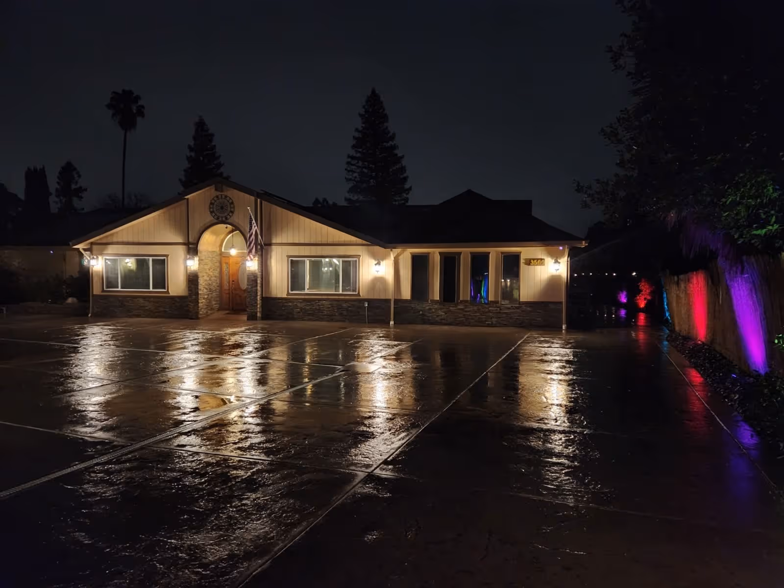 Night view of a single-story building with exterior lights illuminating the entrance and windows. The wet pavement in front reflects the lights, and colorful lights are visible along the right side near a fence and trees.