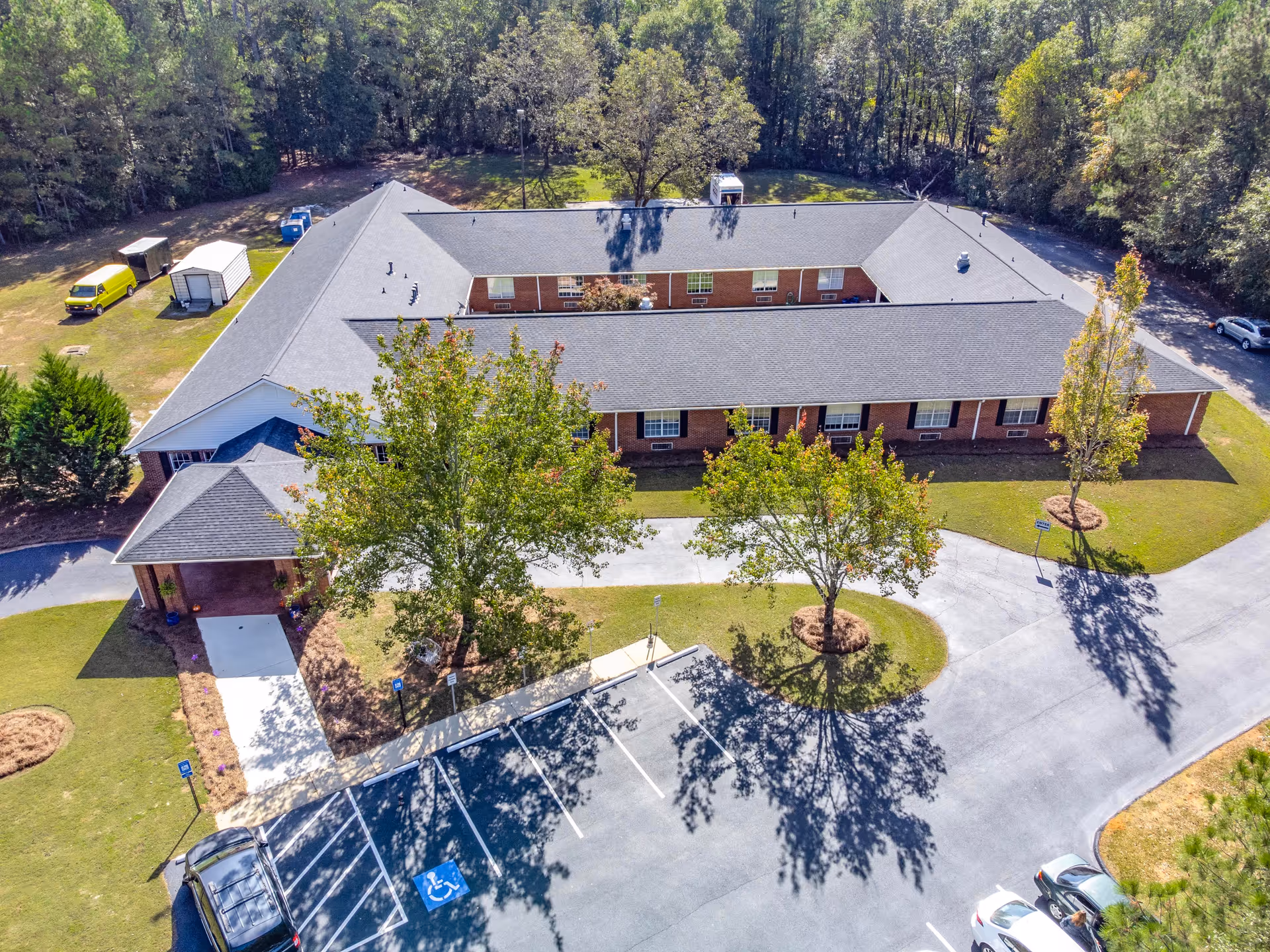 Aerial view of a single-story brick building with a gray roof surrounded by trees and greenery. The building is shaped like a square with an open courtyard in the center. There is a parking lot with several cars and handicap parking spaces in front of the building. The area is well-maintained with a paved driveway and landscaped grass areas.
