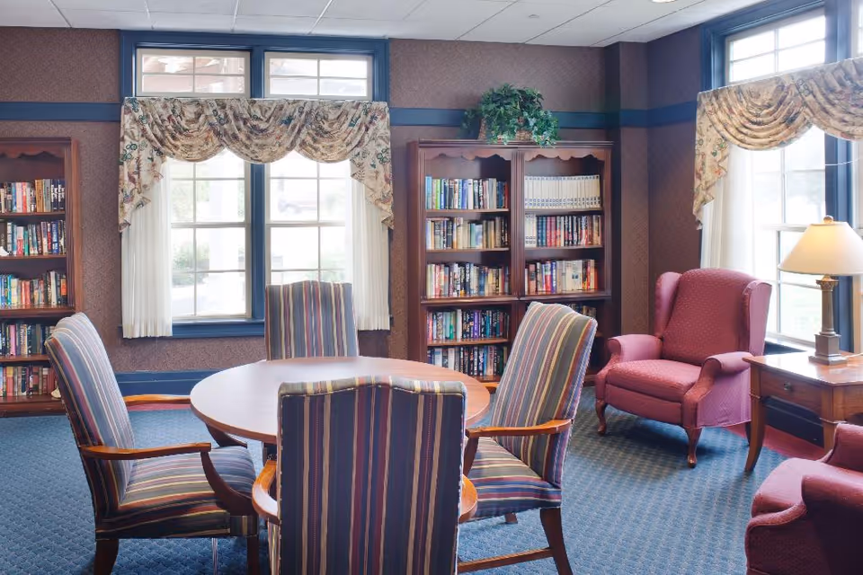 A cozy reading and sitting area with a round wooden table surrounded by four striped upholstered chairs. Behind the table are two large windows with floral valances and white curtains. There are two wooden bookshelves filled with books, a potted plant on top of one bookshelf, and two red upholstered armchairs near a wooden side table with a lamp.