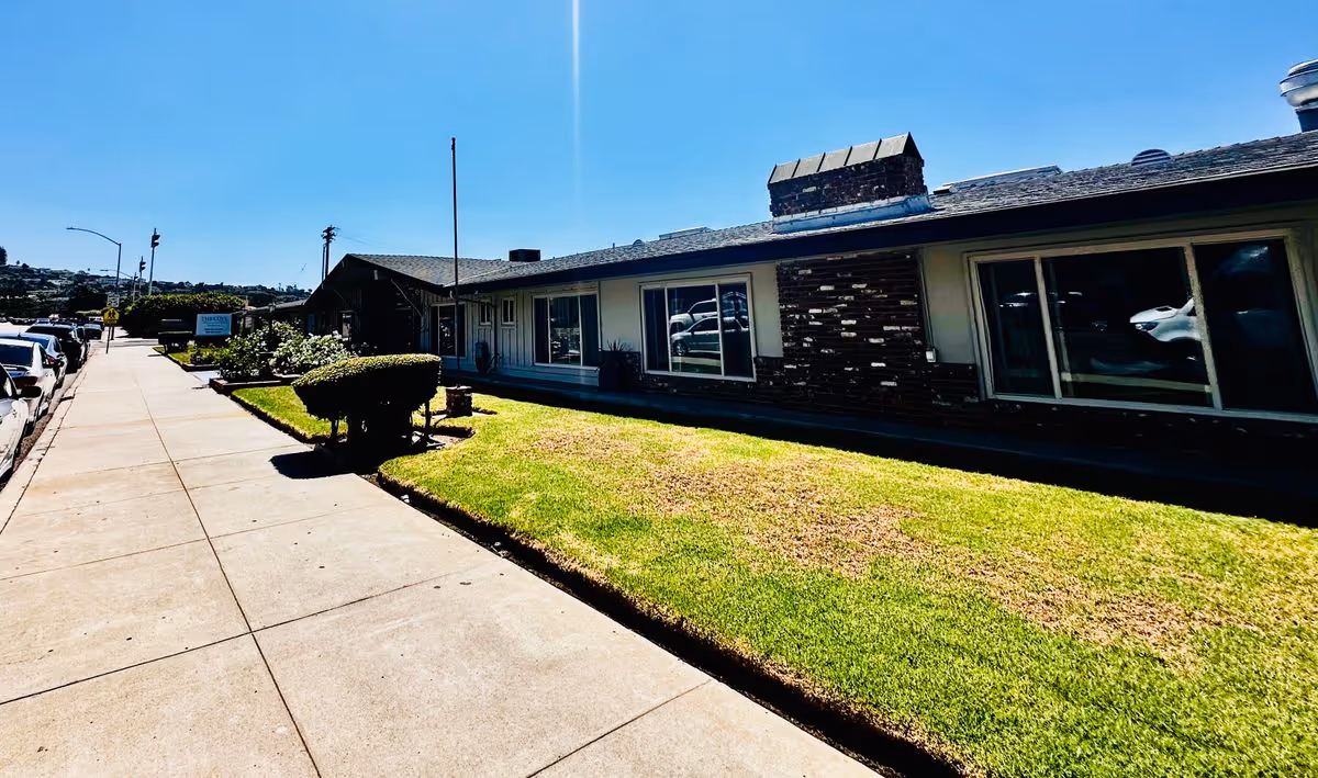 Single-story senior living facility building with a grassy front lawn and sidewalk under a clear blue sky.