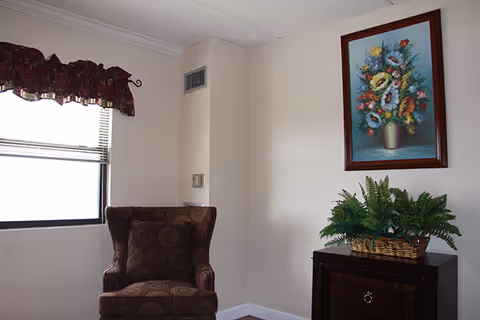 Cozy sitting area with a patterned armchair, a side table holding a potted fern, and a framed floral painting beside a window.