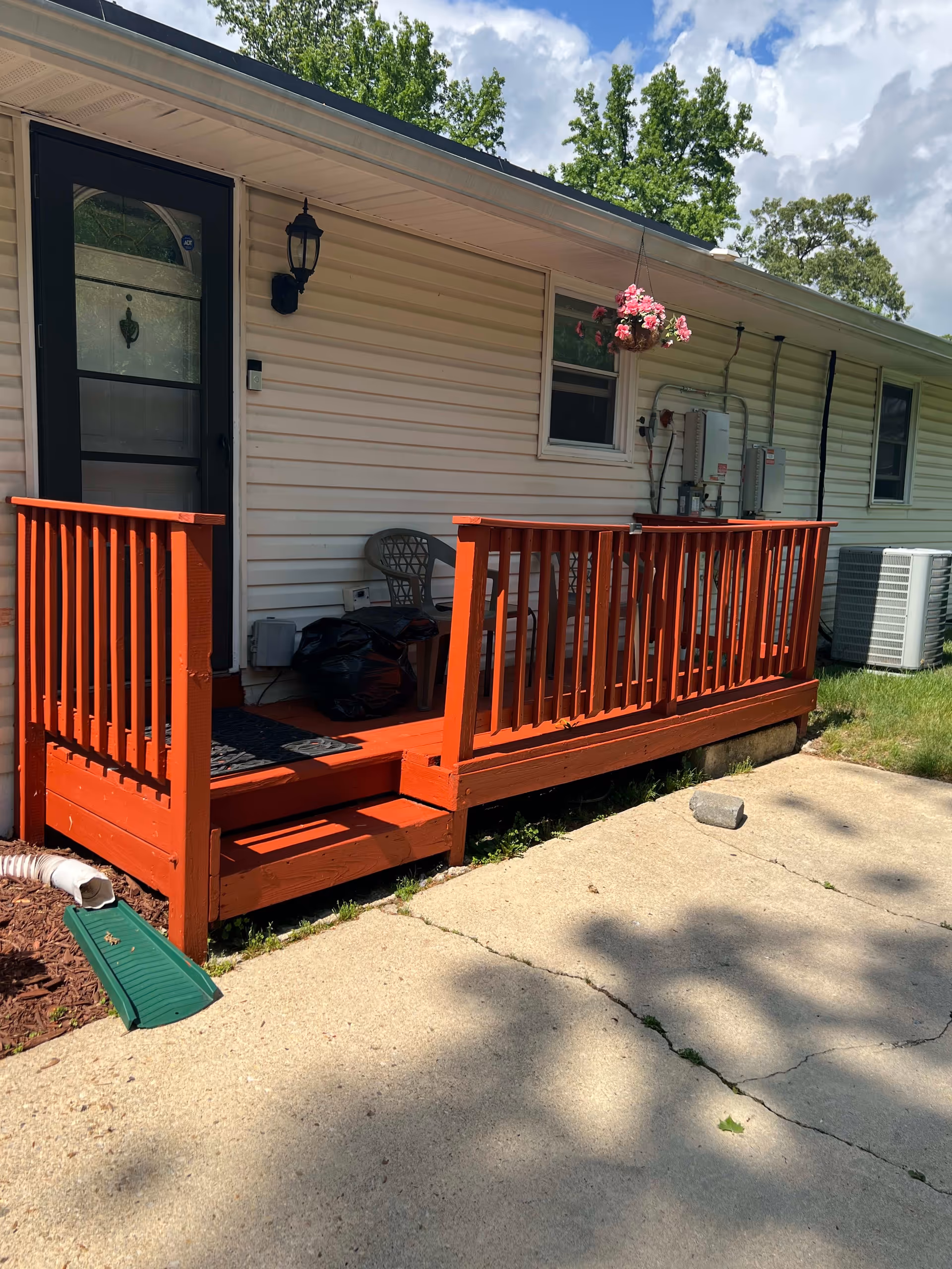 Small red-painted wooden porch with steps and railing attached to a single-story white vinyl-sided house, a black screen door, hanging flower basket, and a concrete walkway.