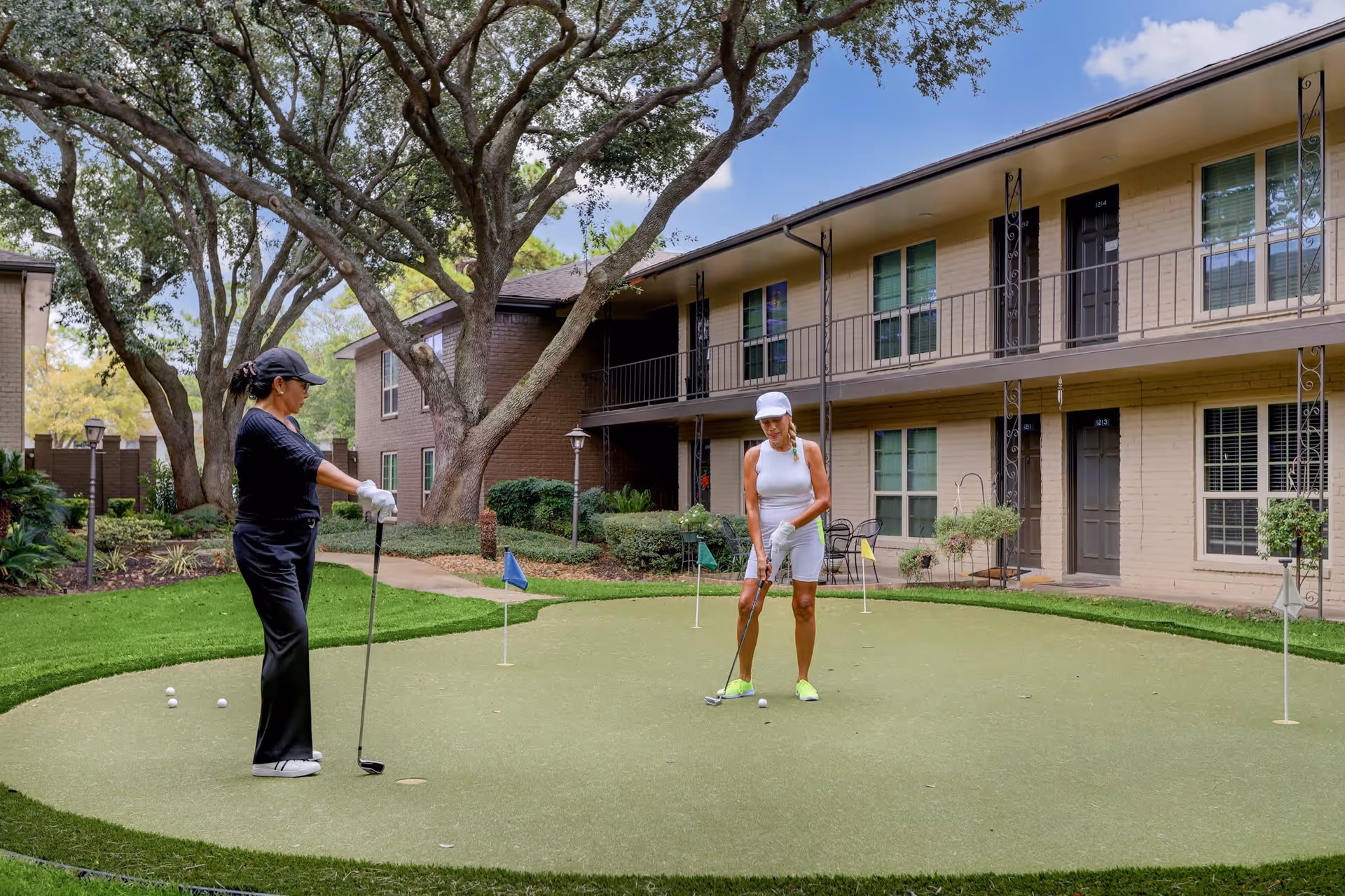 Two women playing golf on a putting green in the courtyard of a senior living facility with two-story beige brick buildings and large trees in the background.