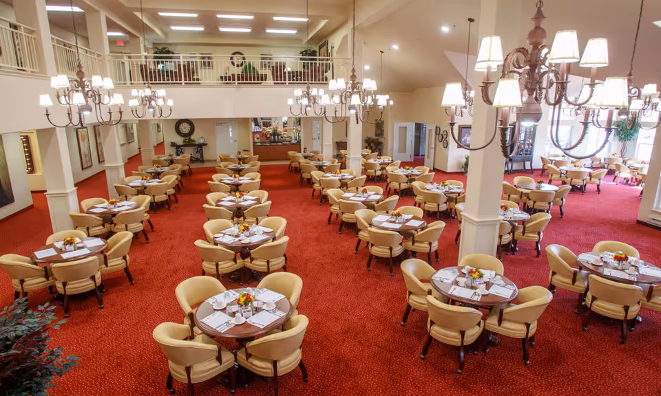Spacious dining room with many round tables set for meals, beige chairs, red carpet, chandeliers, and a mezzanine balcony.