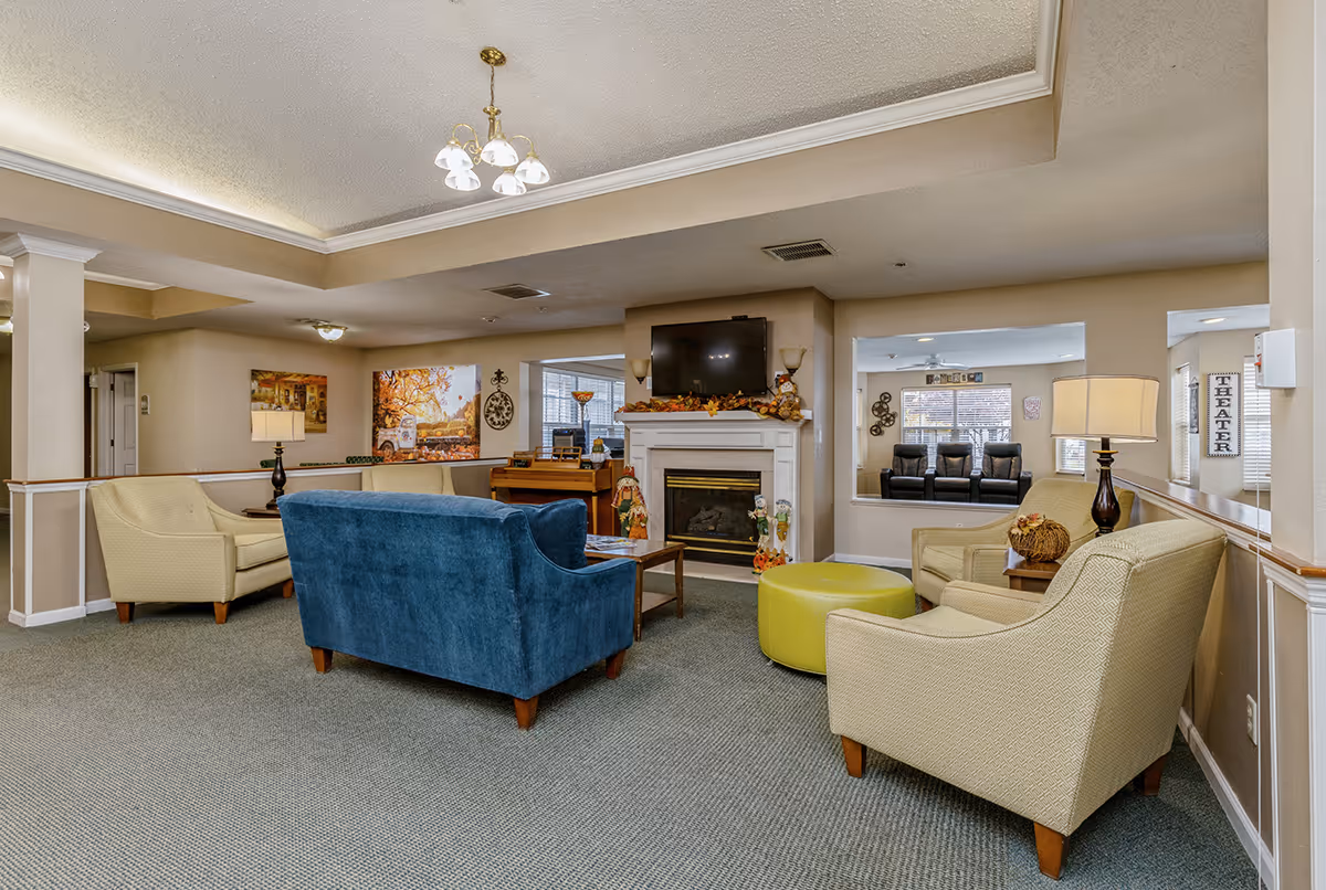 A cozy senior living facility common area with beige walls and carpeted floor. The room features a blue upholstered sofa, two beige armchairs, a green round ottoman, and a wooden coffee table. A white fireplace with fall-themed decorations and a mounted flat-screen TV is centered on one wall. There are two table lamps on side tables next to the armchairs, a chandelier hanging from the ceiling, and a piano in the background. A windowed room labeled 'THEATER' with black recliner chairs is visible through an open wall space.